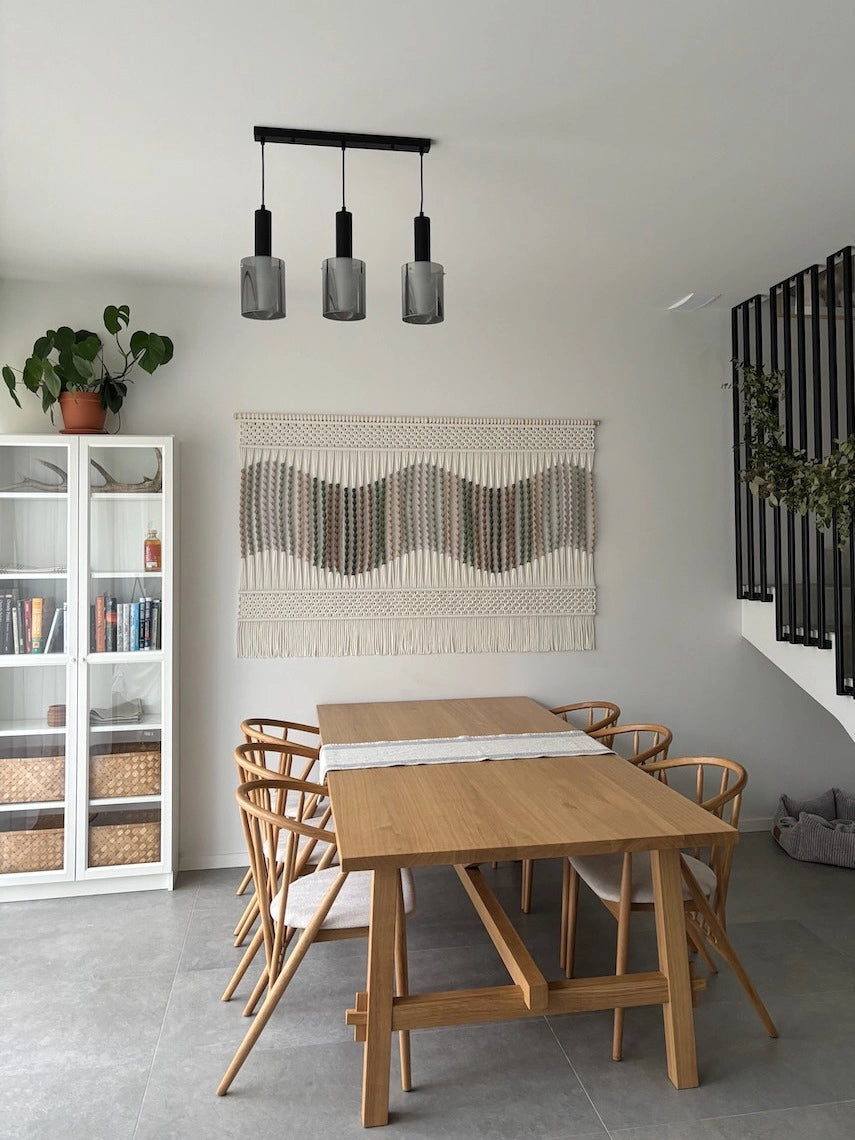 Ręcznie wykonana makrama ścienna w odcieniach beżu zawieszona nad drewnianym stołem w jasnej, przytulnej jadalni w stylu skandynawskim i japandi. Minimalistyczne wnętrze z naturalnym drewnem, prostymi formami, białą witryną i rośliną w tle.